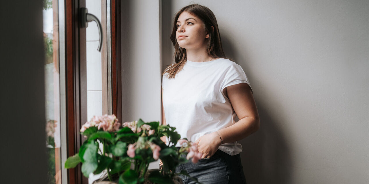 Eine Frau steht an einer Wand angelehnt und schat aus dem Fenster. Sie trägt ein weißes T-Shirt und eine graue Jeans. Auf der Fensterbank stehen Rosen in einem Korb.