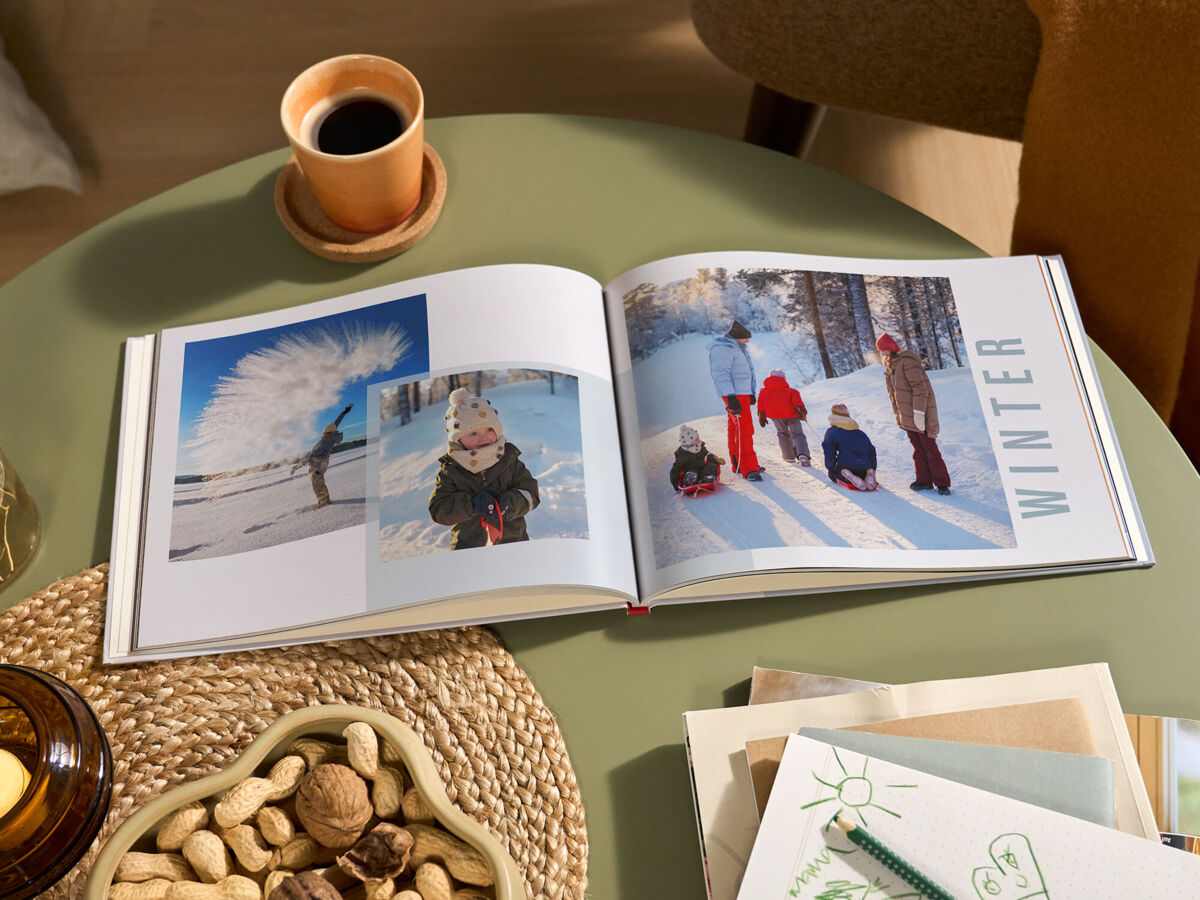 Aufgeschlagenes CEWE FOTOBUCH mit winterlichen Familienfotos im Schnee und der Aufschrift „WINTER“ auf einem grünen Tisch; daneben stehen ei