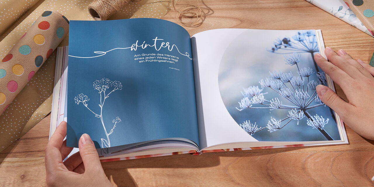 Auf einem Tisch liegt ein aufgeschlagenes CEWE FOTOBUCH. Zwei Hände halten das Fotobuch. Daneben liegen Geschenkpapier und Blumen. Auf der Doppelseite sind eine Nahaufnahme von Pflanzen im Winter mit Frost sowie ein Spruch und die Überschrift „Winter“ zu sehen.