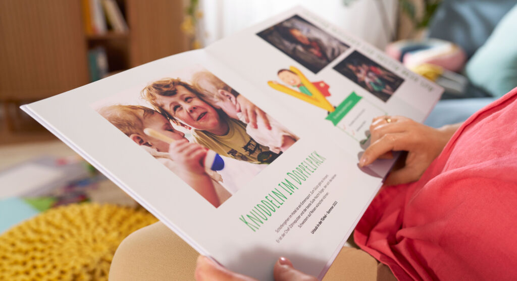 Eine Frau sitzt auf einem Sofa und blättert durch ein CEWE FOTOBUCH, die Doppelseite zeigt Fotos, Text-Elemente und eine Grafik.