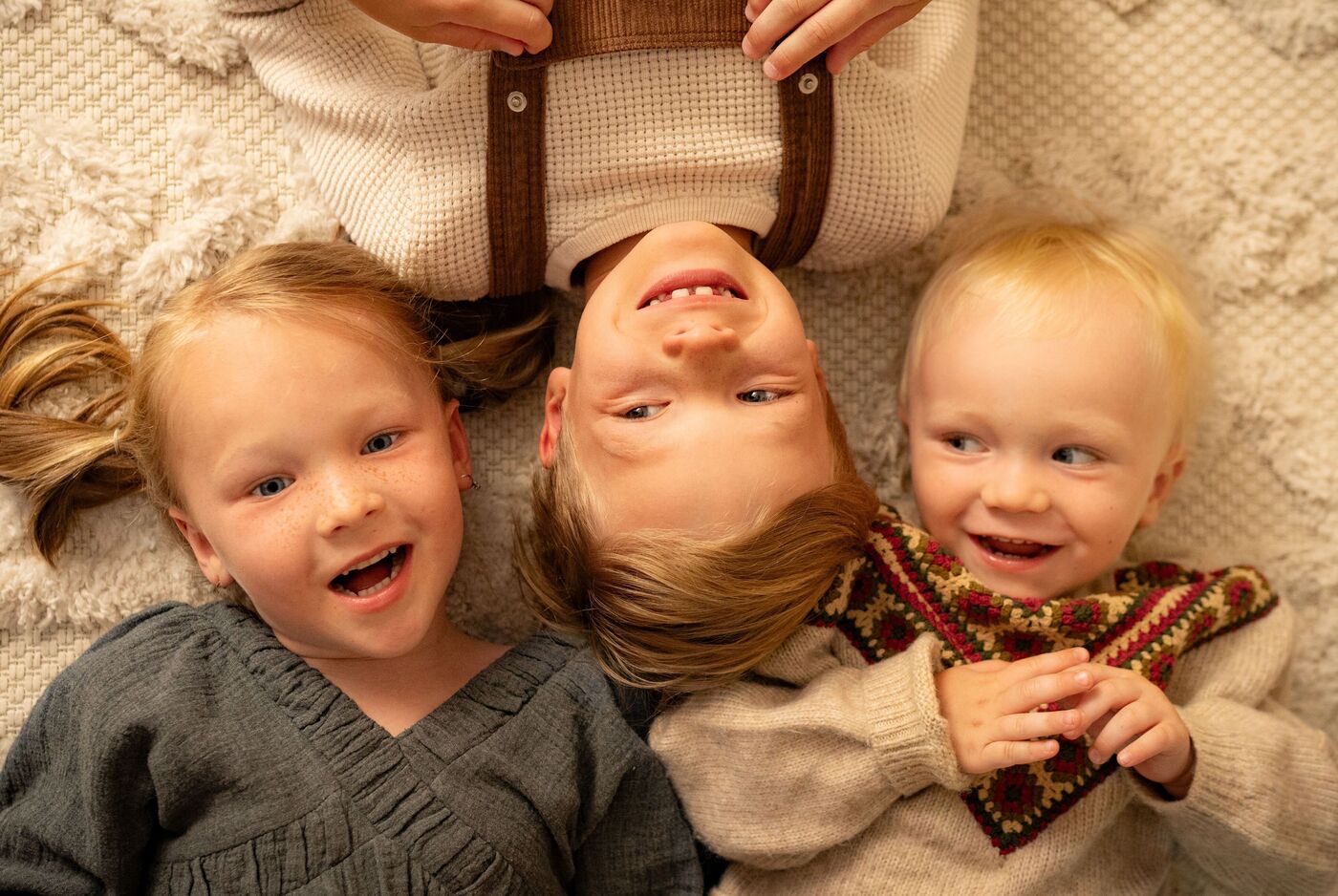 Auf einem hellen Teppich liegen drei Kinder in festlicher Kleidung. Zwei von ihnen blicken lachend nach oben in die Kamera, das dritte Kind schaut fröhlich zu seinem Bruder hinüber, der kopfüber in der Mitte liegt.